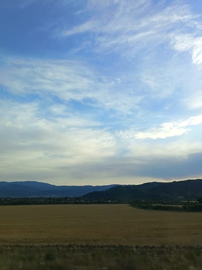 This is a wide, horizontal landscape photograph depicting a rural scene in Skravena, Bulgaria, under a partly cloudy sky. The foreground consists of a dry, golden field, likely harvested wheat or barley, with a strip of darker vegetation running along its edge. In the middle ground, a line of dark green trees borders the field, behind which a small village with scattered buildings is visible nestled at the base of rolling hills. The background is dominated by layers of distant, blue-hued mountains, fading into the horizon. The sky is a bright blue with wispy white clouds stretching across its expanse, some thicker and more opaque closer to the horizon, with the sun's glare faintly visible to the right. The overall impression is of a serene, agricultural landscape on a clear day.