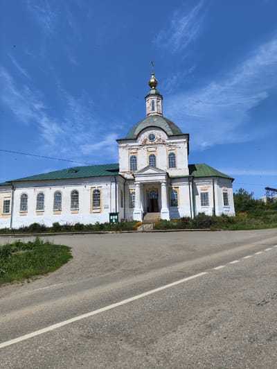 A white, multi-domed church with a green roof stands under a bright blue sky with scattered white clouds. The church has a central tower topped with a golden dome and cross. The building features arched windows with yellow detailing around them. A paved road runs in the foreground, with a grassy verge separating it from the church. The church is situated in the village of Mostovoye, Russia. There are no people visible in the image, and the scene appears calm and undisturbed. Small birds can be seen flying in the sky.
