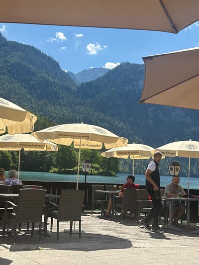 The image depicts an outdoor seating area of a restaurant in Schönau am Königssee, Germany, with a scenic view of a lake and mountains. Several tables with wicker chairs are arranged on a wooden deck. People are seated at some of the tables, enjoying drinks and the view. A waiter, wearing a black vest over a white shirt and black pants, is walking through the seating area. In the background, the Königssee lake is visible, surrounded by lush green, tree-covered mountains under a clear blue sky with scattered white clouds. The restaurant's umbrellas, branded with "Hofbrauhaus Berchtesgaden," provide shade. A lamppost with a traditional design stands near one of the tables. The overall scene conveys a relaxed atmosphere in a picturesque natural setting.