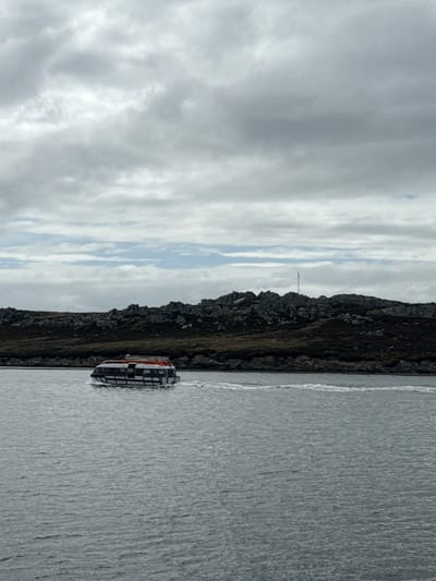 The image depicts a small, enclosed tender boat moving across a body of water in Stanley, Falkland Islands (Islas Malvinas), under an overcast sky. The boat, white with blue stripes and a red roof, travels from left to right, generating a visible white wake behind it. The water surface is dark and rippled. In the background, a dark, rocky landmass rises from the water's edge, characterized by numerous grey boulders and sparse, dark vegetation. A tall, slender mast or antenna is visible on the crest of the landmass, slightly right of the frame's center. The sky is largely covered by grey, layered clouds, with a faint lighter blue strip visible near the horizon line. No individuals are discernible within the boat or on the landmass.