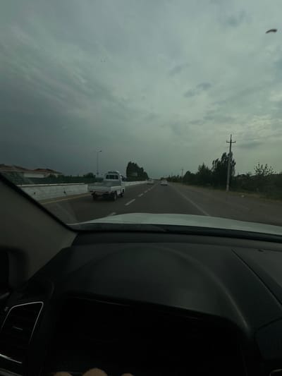 The image shows a view from inside a vehicle, looking out onto a highway. The sky is overcast and appears to be nearing dusk. A white pickup truck is visible on the road, ahead of the vehicle the photo is taken from. There are other cars in the distance on the highway. Along the side of the road, there are trees and utility poles. The dashboard of the vehicle is visible in the foreground. The location is identified as Sergeli, Uzbekistan.