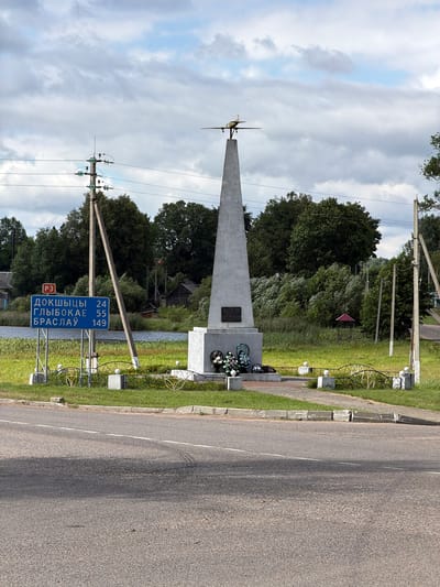 This media file captures an outdoor scene with a monument, road, and sky. The monument is a tall, slender, light-colored obelisk with what appears to be a model airplane or propeller on top. At the base of the monument, there's a plaque and a wreath of artificial flowers. A decorative metal fence surrounds a portion of the monument and adjacent grassy area.
The scene includes a paved road running alongside the monument, with a distinct white line marking its center. There's a grass verge and a sidewalk between the road and the monument. Utility poles with electrical wires are visible, one of which is near the monument and another in the background.
In the background, there are dense trees and some vegetation. A small red-roofed structure is partially visible amongst the trees, suggesting a rural or suburban setting. The sky is partly cloudy, with a mix of light and darker gray clouds, indicating it could be daytime with variable weather.
A prominent blue road sign is mounted on a pole near the monument. It displays Cyrillic text and numbers: "ДОКШЫЦЫ 24", "ГЛЫБОКАЕ 55", and "БРАСЛАУ 149". This sign indicates distances to the towns of Dokshytsy, Hlybokaye, and Braslav, suggesting the location is on a road connecting these places. There's also a red square sign with "РЗ" inside, which is likely a road designation.
There are no people visible in the frame. The overall impression is a roadside memorial or landmark in a natural setting. The lighting suggests it's daytime, likely late morning or afternoon given the shadows. The presence of the monument and the road sign point to this being a significant location along a travel route.