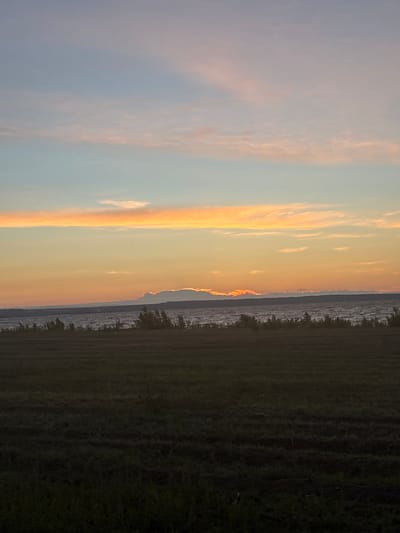 The image depicts a wide landscape scene at sunset. In the foreground, a dark, cultivated field stretches across the lower portion of the frame. Beyond the field, a body of water, likely a river or lake, occupies the middle ground. The water appears choppy, with visible waves. A line of low-lying vegetation and trees runs along the far bank of the water. The sky is a prominent feature, displaying a gradient of colors characteristic of sunset. Soft, horizontal bands of pale pink and orange clouds are visible against a fading blue sky. The sun itself is not directly visible, but its presence is indicated by the warm illumination of the clouds and the horizon. There are no people or specific buildings visible in the image. The scene appears to be in a rural or semi-rural setting. The location is identified as Alexeyevskoye, Russia.