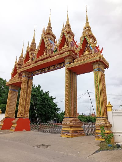 This image captures a grand, ornate temple gate in Bang Kaeo Subdistrict, Thailand. The gate, adorned in vibrant red and gold, features multiple tiered roofs topped with spires. The pillars are intricately decorated with golden patterns, and the lintel displays Thai script. Behind the gate, a dense canopy of green trees is visible, and a collapsible metal fence stretches across the entrance. The sky is overcast, with cloudy formations. The scene suggests a place of worship and cultural significance within the locality.