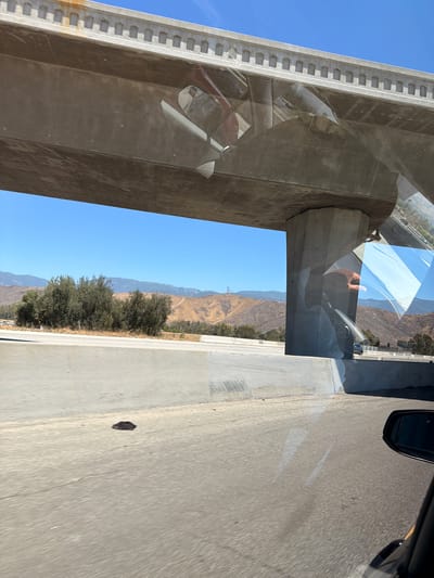 An overpass on a sunny day in San Bernardino, California, is seen from a moving vehicle. The overpass is made of concrete and has a decorative top edge. There are reflections of the vehicle's interior on the overpass's underside. In the distance, dry, rolling hills and mountains are visible, along with some scattered trees and cell towers. A highway with a concrete barrier runs in the foreground, and a dark shadow is cast on the road. The sky is clear and blue.