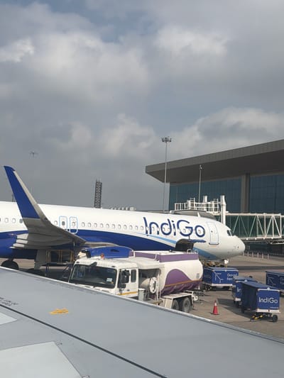 In Vikram Nagar, India, an Indigo Airlines airplane is parked at an airport gate. The aircraft is predominantly white with blue accents and the Indigo logo prominently displayed on its fuselage. A jet bridge extends from the terminal building, a modern structure with a dark grey roof and large glass windows, towards the airplane. In the foreground, the wing of another airplane is visible, with markings that include "DO NOT" and "THIS AREA". On the tarmac, a white refueling truck with a purple stripe is positioned next to the Indigo plane. Several blue cargo carts with the Indigo logo and the text "Ground Force" are also present. The sky is partly cloudy with a mix of white and grey clouds. The overall scene depicts a typical airport environment with aircraft and ground support equipment.