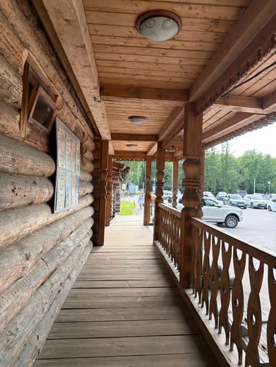 The image captures a traditional wooden structure, possibly a log cabin or a rustic building, in Nevskii Parkleskhoz, Russia. The perspective is from within the covered porch area, looking outwards towards a parking lot with several cars and a line of trees in the background. The architecture is characterized by its extensive use of natural wood, with log walls and intricately carved wooden railings and columns. A long porch with a wooden floor stretches along the side of the building, offering a view of the exterior.

On the porch, a large, framed informational display is laid out, showing what appears to be multiple pages of text and images, possibly menus or informational leaflets. A small, open wooden frame is embedded in the log wall near the display. Two circular light fixtures are visible on the wooden wall, one clearly showing a patterned glass shade.

Beyond the porch, a paved area with parked vehicles is visible, including white and green cars. Some people are seen in the background near the cars and walking further down a set of stairs. The scene suggests a daytime setting, with overcast or bright diffused light, indicating mild weather. The overall impression is one of a rustic, natural environment, likely a tourist destination or a recreational area, given the parking facilities and the informational display. The decorative wooden elements and the use of natural materials create a distinctly regional aesthetic. There is visible text on the informational display, with Russian Cyrillic characters predominantly featured, but the specific content is not legible.