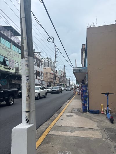 This image depicts an urban street scene in San Juan, Puerto Rico, specifically in the area identified by the "CONTURCE HOSTEL San Juan" sign visible on a building. The perspective is from a sidewalk, looking down a two-lane street under an overcast sky.
In the immediate foreground on the left, a white utility pole, heavily covered with stickers and graffiti, stands prominently. A dark-colored pickup truck is partially visible on the street to the left of the pole. On the right, a light brown multi-story building features a projecting gray awning. Beneath this awning, several blue plastic crates are stacked against the building, and a blue electric scooter, a yellow plastic shovel, and a pink plastic object rest on the concrete sidewalk.
The paved street extends into the distance, bordered by a yellow curb line on the right. Moderate vehicular traffic, including multiple white and dark-colored SUVs, moves away from the camera. Several pedestrians are observed walking away from the camera on the right-side sidewalk. Multi-story buildings, including the "CONTURCE HOSTEL" with green-tinted windows on the left, line both sides of the street. Numerous overhead utility wires and poles are prominent throughout the scene.