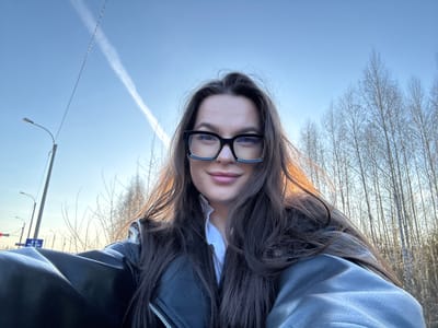 This image is a low-angle portrait of a young woman, originally oriented upside down. When viewed upright, she is seen looking directly towards the viewer, framed by her long, dark brown hair which cascades around her face and shoulders. She wears thick-framed black glasses and her lips have a natural, light pink hue. Her attire includes a black leather jacket over what appears to be a white shirt, visible at the collar. Her expression is neutral and composed.

The background on her left side features a dense collection of slender, bare trees, characteristic of late autumn, winter, or early spring. On her right side, the backdrop opens up to a wide expanse of clear, light blue sky, marked by a distinct white contrail from an airplane stretching diagonally. Urban infrastructure elements are visible in the distance, including utility poles, a street light with a curved arm, and a blue rectangular road sign displaying two white arrows—one pointing left and one pointing right, stacked vertically. There is no discernible text on the sign.

The lighting is bright and natural, indicating daytime conditions. The scene suggests an outdoor setting, possibly along a road or in a natural area bordering urban development, located in Novoe Mart'yanovo, Russia.