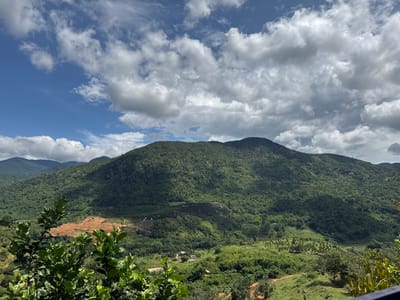 The image captures a scenic vista of rolling green mountains under a partially cloudy blue sky. In the foreground, lush green foliage is visible, framing the expansive landscape. The midground features a cleared, reddish-brown patch of land on a hillside, likely for agricultural or development purposes, contrasting with the dense, verdant vegetation that covers the rest of the terrain. Scattered amidst the greenery, several small buildings and structures can be observed, suggesting the presence of a rural settlement. The background is dominated by layers of mountains receding into the distance, their slopes uniformly covered in thick vegetation. The scene depicts a natural environment with evidence of human presence in the form of cleared land and buildings. The location is identified as La Dạ, Vietnam.