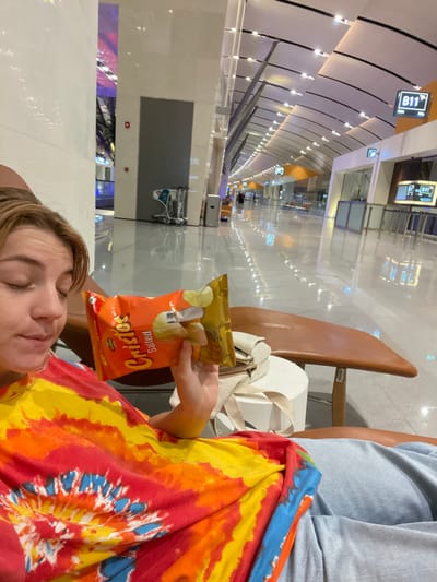 A fair-skinned individual is depicted seated in a brown lounge chair within Muscat International Airport (MCT), located in Seeb, Muscat Governorate, Oman. This identification is supported by the visible "B11 Gate" signage and the characteristic modern architecture of the airport terminal.

The individual, appearing to be in a relaxed state with partially closed eyes and a slightly open mouth, wears a vibrant tie-dye t-shirt featuring red, orange, yellow, and blue patterns, along with light grey pants. In their right hand, they hold an open, orange-colored bag of "Cristos Salted" potato chips. A light-colored shoulder bag rests on a small white cylindrical table positioned next to the lounge chair.

The airport environment features a highly polished, reflective light-colored floor and a distinctive high, vaulted ceiling with integrated lighting. In the background, a long, well-lit terminal corridor extends, populated by several indistinct human figures, additional seating, a metal luggage trolley, and a grey waste bin. Multiple digital signs displaying "B11 Gate" are visible further down the concourse on the right.