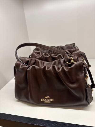 The image shows a brown Coach purse with a gathered or ruched design. The purse is positioned against a white surface, likely a shelf or wall, with a dark-colored trim or frame visible to the right. The purse features a gold Coach logo and hardware. The location is in the city of Sunrise, United States of America.