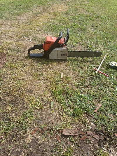 A Stihl chainsaw lies on its side on a grassy area, with its chain bar facing to the right. The chainsaw is orange and silver. To the right of the chainsaw, a yellow and black measuring tape, a red pencil, and a wooden ruler are placed on the ground. The ground is covered in dry grass and patches of green grass, with some scattered dirt and small pieces of bark. The image appears to be taken outdoors on a sunny day, likely in a yard or park within Удзелаўскі сельскі Савет, Belarus.