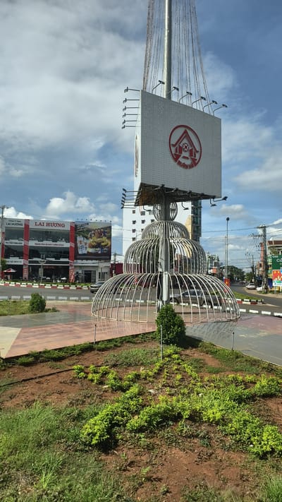 The image shows a street scene in Buon Ho, Vietnam. In the foreground, there is a landscaped area with red soil and green vegetation, leading to a circular metal sculpture. Behind the sculpture, a large white billboard is visible, featuring a red circular logo and text. To the left of the billboard, a multi-story building with a red and white facade is seen, with the words "LAI HUONG" in large lettering and other signs. Further in the background, there are more buildings, a street with traffic, and power lines against a cloudy blue sky. The overall impression is of a commercial area on a bright day.