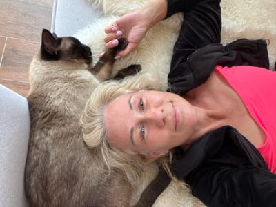 In the city of Kerro, Russia, a woman with blonde hair lies on a fluffy rug next to a Siamese cat. The woman is wearing a pink tank top and a black zip-up jacket. Her hand is gently resting on the cat's paw. The cat is facing away from the camera, with its head partially visible. The background shows a wooden floor and a light-colored sofa.