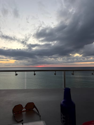 The image captures a view from what appears to be a balcony. The left side of the image displays a sky filled with grey and white clouds. There's a hint of red and yellow sunlight peeking through. A glass railing with metal supports separates the sky and sea from a grey, flat surface. On this surface, there's a dark blue bottle laying on its side, and a pair of brown tortoise-shell sunglasses. A white object is also visible beside the sunglasses. The location is likely in San Miguel de Cozumel, Mexico, as the presence of the ocean would indicate.