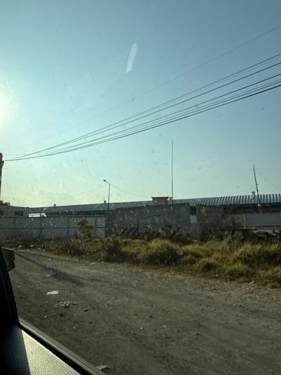 A view from inside a vehicle, likely a car. Power lines are visible against a light blue sky. On the right, a building runs parallel to a dirt or gravel road. Grass grows along the building's edge. The windshield appears to have water spots. The interior of the car is visible in the bottom right corner.