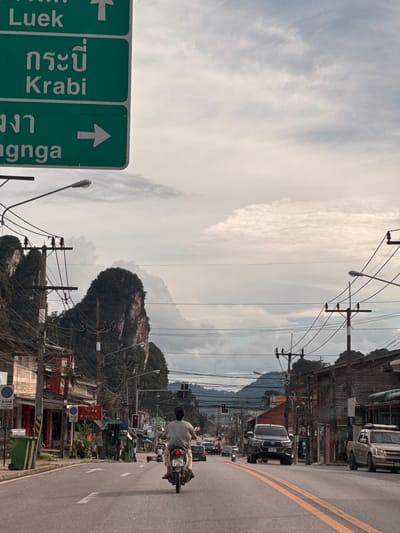 The image depicts a street scene in Ao Luek, Thailand, characterized by a road with traffic and buildings lining its sides. A prominent green road sign with white text indicates directions to "Luek," "Krabi," and "Nganga," with arrows showing the respective routes. A person is riding a scooter in the foreground, heading away from the viewer. Other vehicles, including cars and motorcycles, are visible on the road. The background features dramatic karst mountains characteristic of the region, partially obscured by an overcast sky. Electrical poles and wires are abundant, contributing to the urban infrastructure of the area. The buildings on either side of the road appear to be a mix of commercial establishments. The overall atmosphere suggests a typical day in a Thai provincial town, with a blend of natural scenery and local life.