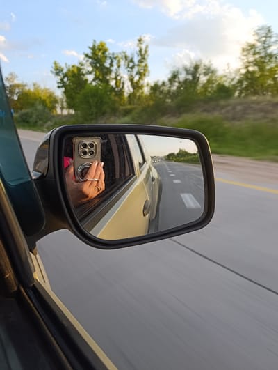 A side-view mirror reflects a person holding a smartphone while in a moving vehicle on a highway. The phone has a square camera bump with four lenses and a circular ring on the back. The person is wearing a ring on their right hand. The highway has white lines marking the lanes and traffic can be seen in the distance. The surrounding environment consists of trees and a clear sky with clouds. The image was taken in Rassvet, Kazakhstan.
