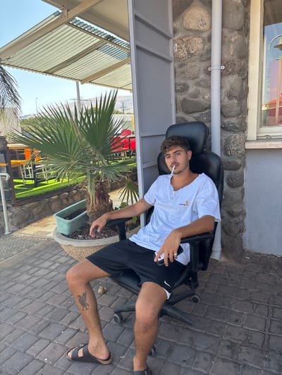 A young adult male is seated in a black office chair outdoors on a paved patio in the Galil Elyon Regional Council, Israel. He wears a light grey, short-sleeved Carhartt t-shirt, black athletic shorts with white trim, and dark brown slip-on sandals. A cigarette is positioned on his lower lip. He holds a black smartphone in his left hand, while his right arm rests on his right leg. A visible tattoo, appearing to be a stylized bird, is on his left lower leg. The ground beneath him is covered in dark grey paver stones. To his right, a building wall constructed from natural, irregularly shaped stones is visible, featuring a white vertical pipe and a window. To his left, a large palm tree is planted in a concrete planter, with a smaller green plastic planter behind it. Beyond the palm tree, an outdoor seating area with red chairs, orange tables, and green artificial turf is present. The background reveals distant hills under a clear blue sky, with electrical poles and lines. Above the scene, a white corrugated metal awning provides shade. The lighting indicates bright daylight conditions.