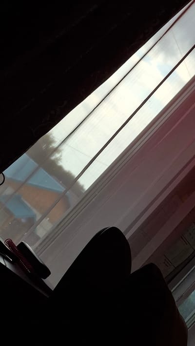 A dimly lit interior scene is captured from a low angle, looking towards a window covered by sheer, vertically striped curtains. Sunlight streams through the translucent fabric, creating bright, elongated rectangles of light and casting shadows from the room's elements.
In the foreground, to the left, a dark, heavy curtain is visible, obscuring much of the room and creating a strong contrast with the illuminated window. To the right, the dark silhouette of a chair or sofa is prominent, its form softened by the low light. Resting on a surface in the lower right corner of the frame is a notebook with a red spiral binding, partially obscured by the darkness.
Through the sheer curtains, the exterior environment is faintly visible. Hints of trees and the roofs of buildings with a blue and orange color scheme suggest a residential area. The quality of light, with its soft diffusion and long shadows, indicates it is likely late afternoon or early evening. The overall mood is quiet and contemplative, with the play of light and shadow creating a sense of depth and mystery. No text is visible in the image. The setting appears to be indoors, possibly a living room or study, with a view of the outside world.
