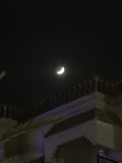 The image shows a crescent moon in a dark sky. The moon is on the left side of the frame. On the right, there's a building with ornate architectural details, possibly made of brick and stone. A palm frond extends from the bottom right corner towards the building. The scene appears to be at night in Skhirat, Morocco.