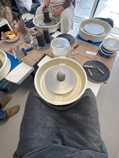 An indoor pottery workshop in Mafra, Portugal, is depicted from a first-person perspective, looking over a dark gray blanket covering the observer's lap. In the immediate foreground, a cream-colored pottery wheel basin holds a metallic wheel head centered with a spherical lump of light gray clay. To the right of this wheel, on a wooden surface, a black circular bat displays three wooden-handled wire cutting tools. The observer's feet, clad in brown boots and blue pants, are visible on a light gray tiled floor to the lower left.
The midground features a wooden tabletop serving as a shared workspace, laden with pottery tools and materials. These include several cylindrical clay forms, some resting on circular bats, and two white plastic buckets, one partially filled. Various hand tools such as a metal scraper with a blue handle, a wooden rib, a metal ruler, carving tools, and sponges are scattered across the surface. A bowl containing colorful sponges and an orange container, along with a container holding numerous modeling tools, are also present.
In the background, a second cream-colored pottery wheel basin is visible on the right. To the upper left, another individual is seated and actively engaged with a third pottery wheel, shaping a cylindrical piece of clay with both hands. This person wears light-colored clothing over a brown long-sleeved shirt. A large window or glass door in the distant background allows natural light into the space, which features a light gray tiled floor throughout. The scene is an active and prepared environment for ceramic production.
