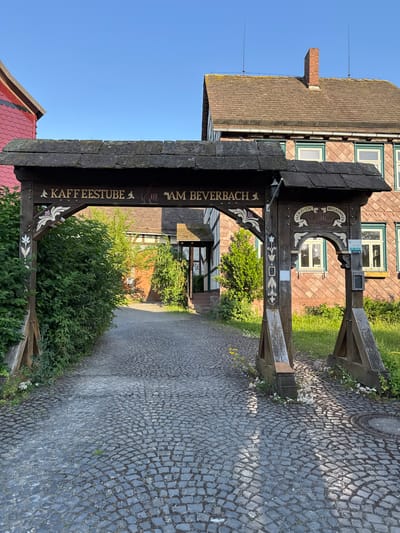 This is a vertical, eye-level, outdoor, daytime photograph of an ornate wooden archway leading into a paved courtyard. The archway is decorated with carvings and painted details. Visible text on the archway reads "Kaffeesstube" and "am Beverbach," indicating the name of a coffee house located by the Beverbach. Above, carved into a decorative curved section, is the word "WILLKOMMEN" in German, meaning "Welcome."

Behind the archway, a traditional German building with a red tiled roof and dark wood trim is visible. The building has several windows with light-colored frames. Beyond this building, the paved courtyard extends, bordered by greenery and more buildings in the distance. The cobblestone paving is a prominent feature of the ground. The sky is clear and blue, suggesting fair weather. The scene appears peaceful and inviting, typical of a rural German village setting. The time of day is likely late morning or early afternoon, given the strong sunlight and shadows cast.