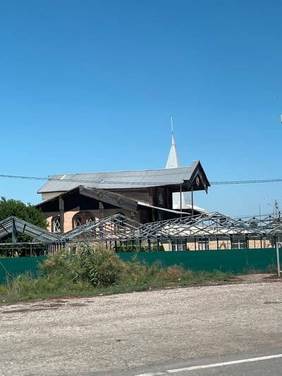 This is a photograph taken on a clear day, showing a building with a distinctive architectural style in Il'inka, Russia. The building appears to be constructed primarily of wood, with a light-colored brick base visible in parts. It features a prominent, multi-pitched roof with a tall, pointed spire extending upwards. Several arched windows with intricate latticework are visible on the building's facade. In the foreground, there is a green metal fence, behind which a structure made of metal framing and possibly some unfinished roofing material is situated. Beyond the fence, dense green foliage and dry, brown vegetation are present. A rough, gray, unpaved surface, possibly a road or parking area, occupies the bottom portion of the image. Power lines are visible stretching across the sky in the upper portion of the image. The overall impression is of a religious or historical structure in a semi-rural or developing area. There are no people visible in the image.