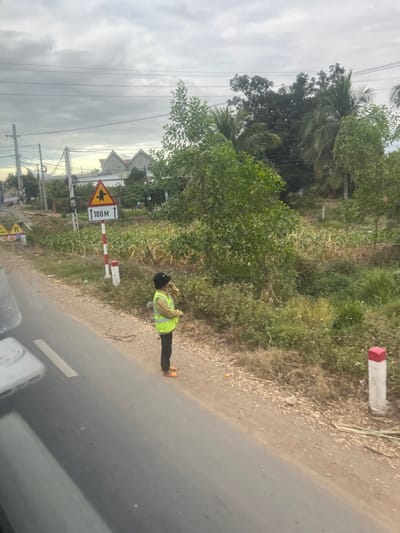 The image depicts a roadside scene in Ninh Tây, Vietnam, under an overcast sky. A single individual stands on the unpaved dirt shoulder of an asphalt road that runs along the lower left quadrant of the frame. The person is dressed in a black bucket hat, a light-colored long-sleeved shirt, a neon yellow-green reflective safety vest, dark trousers, and orange footwear. They hold an object to their mouth or chin with their right hand, appearing to consume something, while facing generally towards the right.
To the left of the individual, a white signpost with a red top supports a triangular yellow and black warning sign featuring a pedestrian or child figure. Below this, a rectangular white sign indicates "100 M" with an upward arrow. Further right along the shoulder, another white post with a red top marks the roadside.
Beyond the shoulder, the environment transitions into dense green vegetation, including tall grasses, shrubs, and small trees. In the mid-ground, cultivated fields with row crops are visible, leading to several houses with gabled roofs in the distant background. Utility poles and overhead power lines are present on the left side of the frame. A partial view of a white vehicle component is visible in the lower left corner, suggesting the image was captured from within a vehicle.