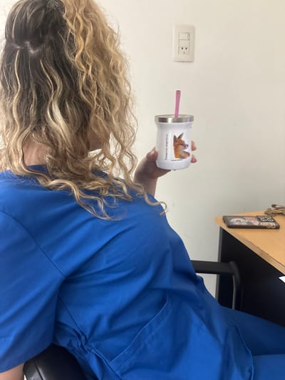 A person in blue scrubs sits with their head tilted, revealing long, wavy, blonde hair. They hold a white tumbler with a fox image and the words "Patagonia Argentina" on it, and a pink straw sticks out of the tumbler. A light switch is visible on the wall. A portion of a desk and a cell phone can be seen in the upper portion of the frame.