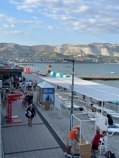 This is an outdoor, elevated, eye-level shot of a crowded beach area with a mountain range and a harbor in the background. In the foreground, there is a paved walkway with several people walking and standing. To the right of the walkway, there is a beach with numerous white lounge chairs, some occupied by people, and a white canopy. A small kiosk with white framing and a white fabric roof is in the mid-right, with people interacting around it. Further to the right, there are more beachgoers and lounge chairs.
On the left side of the image, there is a red structure with the word "BOXER" in white letters. Adjacent to it is a food or drink stand with the word "ТИР" in green letters. Several people are gathered around these structures.
The beach itself is covered in grey pebbles. People are seen swimming and wading in the calm, blue water. A concrete pier extends into the water in the mid-ground, with a few individuals walking on it. In the distance, a large mountain range with terraced slopes and some industrial structures, including red and white smokestacks, is visible. A harbor with cranes and ships is situated at the base of the mountains. The sky is light blue with scattered white clouds.
The overall scene depicts a lively beach resort atmosphere on a sunny day.