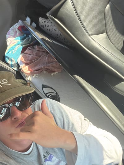 A person wearing sunglasses and a hat is seen in the front seat of a car, next to a surfboard. The surfboard is resting on the open car door, which is filled with towels. The car's leather seat is visible in the upper left. The person's hand is visible, pointing downward. The photo appears to have been taken from a high angle.