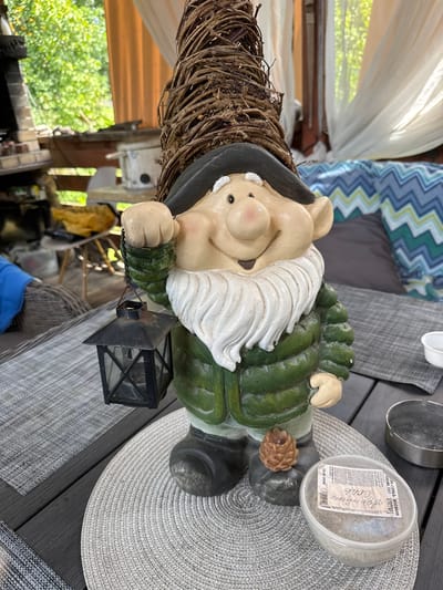 In the city of Yesipovo, Russia, a garden gnome with a twig hat stands on a wooden table. The gnome is dressed in a green jacket and black boots, and it holds a black lantern in its left hand. A small pinecone-shaped candle sits on the gnome's right boot. Next to the gnome, a clear plastic container with a label contains sand or small stones. An ashtray is visible on the table to the right of the container. In the background, there are outdoor seating areas with cushions and curtains, and lush green trees. The scene appears to be an outdoor patio or balcony.