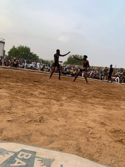 The image shows two men wrestling in a sandy arena, located in the city of Sokoto, Nigeria, at the coordinates 13.049 latitude, 5.238 longitude. The men are shirtless and wearing traditional wrestling attire. One man is holding a stick in his right hand, while the other is striking with his left hand. A large crowd of spectators is seated around the perimeter of the arena. The ground is sandy, and there are some bags in the foreground. The sky is overcast. The men are facing each other, their stances are aggressive. The crowd is watching intently, some clapping and cheering. The overall scene is a traditional wrestling match.