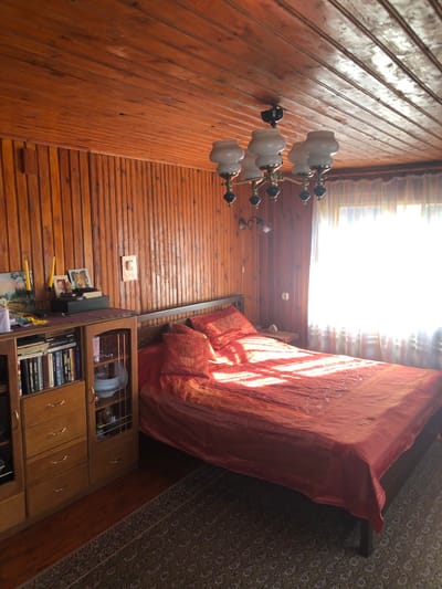 This image depicts an interior room, likely a bedroom or living space, located in Сапоцкінскі сельскі Савет, Belarus. The room features walls and a ceiling entirely clad in horizontal and vertical light-brown stained wood paneling. A large bed with a dark frame and headboard occupies the right half of the scene, covered by a reddish-orange duvet and two matching pillows. Direct sunlight streams through a large window on the far right, partially obscured by sheer, light-colored curtains, casting distinct patterns of light and shadow across the bed. A small wooden nightstand is positioned to the right of the bed.
A five-arm chandelier with white glass shades and dark accents hangs from the wooden ceiling in the upper center. Below it, on the paneled wall, is a two-bulb wall sconce with decorative metalwork. To the left, a tall, multi-section wooden furniture unit stands against the wall. Its upper left section functions as a bookcase filled with numerous books, while below are several drawers with brass-colored pulls. The lower right section is a glass-fronted cabinet displaying plates and other items. The top surface of this unit holds a decorative candelabra with two yellow candles, several framed photographs, and a painting leaning against the wall. A patterned rug covers a portion of the wooden floor in the foreground. No individuals are present in the scene.