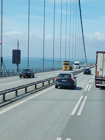 This media file captures a view from a vehicle traveling on a suspension bridge, likely the Great Belt Bridge in Korsør, Denmark. The scene depicts a bright, sunny day with a clear blue sky and scattered white clouds. The bridge spans a wide expanse of calm, blue water.
The primary subjects are vehicles on the bridge. In the foreground, a dark blue SUV is visible. Further ahead, a black car is driving in the same lane. In the opposite lane, a bright yellow truck is prominent, followed by a white truck. Another black car is in the distance. A large white semi-trailer truck is also visible in the right lane of the foreground.
Notable objects include the suspension cables of the bridge, which run vertically through the frame, and the bridge's railings. On the left side of the road, a tall structure with a large black rectangular screen is mounted on a pole, possibly an electronic sign. The overall impression is one of orderly traffic flow on a well-maintained bridge.
The time of day appears to be midday, given the bright sunlight and shadows cast by the bridge's railings. The weather is pleasant, suggesting a good day for travel. The presence of multiple large trucks and cars indicates ongoing traffic. The view of the distant bridge structure further emphasizes the scale and grandeur of the crossing. No people are clearly discernible in the image.
Visible text includes license plates on the vehicles. The black SUV has a license plate that appears to read "FHH 758". The black car in the foreground has a license plate that reads "AX 61197". The yellow truck has a license plate that is partially visible. The white semi-trailer truck has some text and symbols on its rear.