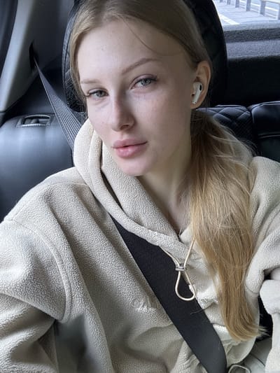 A young woman with blonde hair tied in a ponytail, wearing a light-colored fleece hoodie and a seatbelt, is pictured from the chest up. She has on white wireless earbuds and a necklace. The image appears to be taken inside a car, with the dark interior of the vehicle visible in the background. The scene is indoors, likely in a vehicle. The location is specified as Cheremsha, Russia.