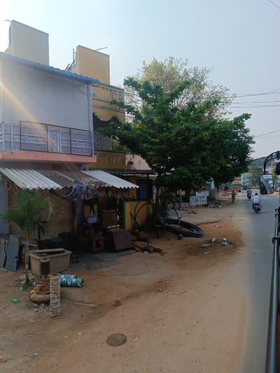 The image depicts a street scene in Kothacheruvu, India. The foreground shows a dirt and gravel shoulder adjacent to a paved road. A collection of objects, including metal pipes, tanks, and machinery parts, are scattered along the shoulder. A small palm tree grows in a pot near these objects. A corrugated metal awning covers a storefront. The facade of the building above the awning is pale gray, with a patterned balcony railing. A tree with dense green foliage obscures part of a building with yellow paint and detailed trim. In the background, a person rides a scooter down the road. Another person rides a motorcycle. A yellow school bus with the word "NAVATA" printed on the front is visible. Power lines stretch across the sky. The view is taken from inside a vehicle with part of the frame visible on the right side of the image.