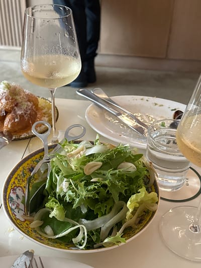 A table setting features a salad bowl filled with lettuce and almonds, accompanied by salad tongs. Nearby, a wine glass is partially filled with liquid, and a half-eaten plate suggests a previous meal. Another glass and a bottle are also present. An additional plate of food is also within view.