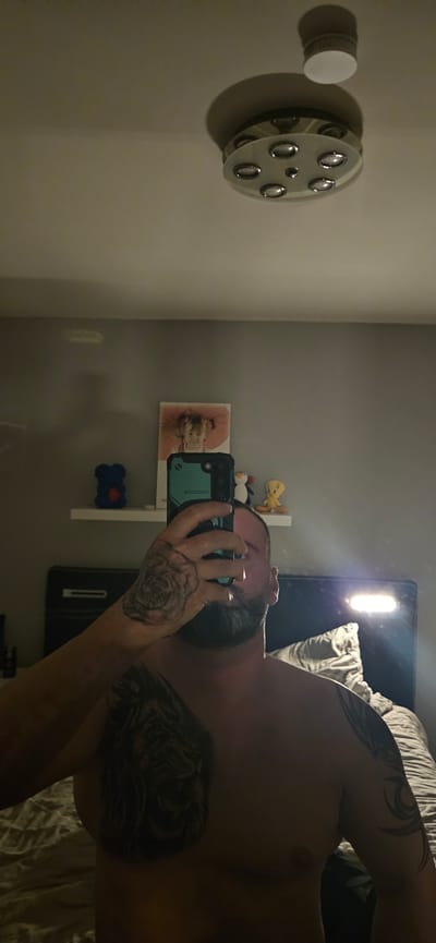 A man with tattoos on his arm and chest is taking a selfie in front of a mirror. A light fixture is on the wall and a shelf with toys and a framed photo is on the wall behind him. The location is likely a bedroom in a home in Gütersloh, Germany.