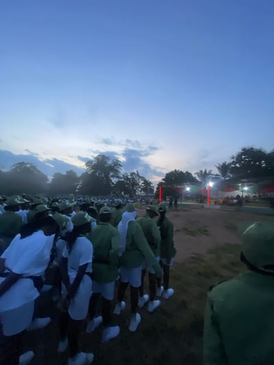 The image depicts a large gathering of young people, appearing to be participants in a youth service program, standing in an outdoor field under a twilight sky. The majority of individuals are wearing matching green uniforms with white accents and green caps. Some are also wearing white t-shirts and shorts. The scene suggests an organized event, possibly an assembly or a parade, given the organized rows of people. In the background, there are trees, tents, and lights, indicating an event space with some infrastructure. The ground is a dirt or sandy surface. The lighting suggests it is either early morning or late evening. The specific location is identified as Orun Ekiti, Nigeria.