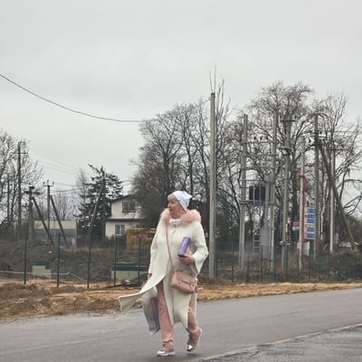 The image shows a woman walking along a road in Skotnoe, Russia. She is wearing a white coat with pink fur trim, pink pants, and pink shoes. She is carrying a pink purse and a box labeled "DéLisse". The road is wet and there are bare trees and utility poles in the background. A house and a green fence are visible in the middle ground. The sky is overcast.