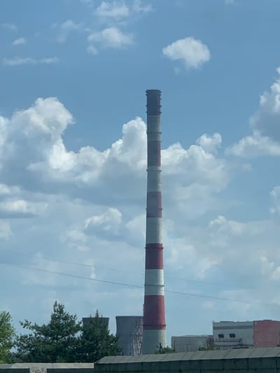 The image shows a tall, red and white striped chimney against a blue sky with white clouds. There are two smaller, gray cooling towers visible in the background, behind the chimney. The chimney is located near a industrial complex. The surrounding area is characterized by a low wall and green trees in the foreground. The scene was captured on a sunny day with visible power lines overhead. The location is Podvirky, Ukraine, which is near a major industrial zone.