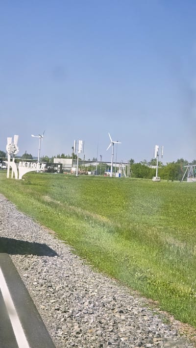 The image depicts a roadside view of a grassy field with several wind turbines and a large white sculpture bearing the text "АКТЮБИНСКИЙ". The sculpture appears to be a stylized representation of a person or figures. In the background, there are some industrial buildings, trees, and more wind turbines of varying designs. A gravel road with a white line runs along the left edge of the frame, and a shadow is cast across it. The sky is clear and blue. The scene appears to be an open area, possibly on the outskirts of a town or industrial zone. The "АКТЮБИНСКИЙ" text suggests the location is related to the city of Aktobe in Kazakhstan or a similarly named settlement.