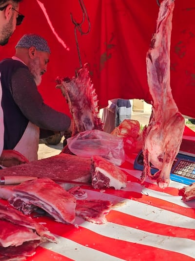 A butcher, donned in a skullcap and grey beard, stands amidst hanging meat cuts at an outdoor stall. Red and white striped material forms the backdrop, while various cuts of raw meat are prominently displayed. A weighing scale is visible, suggesting a commercial transaction. A customer wearing sunglasses is visible on the bottom left. The scene captures a traditional butcher shop setting, filled with fresh meat and the tools of the trade.