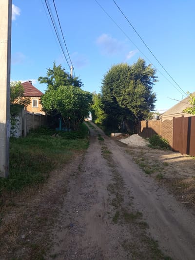 The image depicts a rural or semi-rural street scene under a clear blue sky with scattered clouds, likely in the city of Vilshany, Ukraine. A dirt road, showing tire tracks and scattered gravel, leads upwards and away from the viewer, flanked by vegetation and fences. On the left, a concrete utility pole stands prominently in the foreground. Adjacent to the road is a grassy area with overgrown weeds and dry grass. Further down the left side of the road, a brick house with a brown tiled roof and a single window is visible behind a concrete fence. Several trees with dense green foliage are present, with one particularly large tree dominating the right side of the road, casting a shadow. On the right side of the road, a beige brick fence with a decorative pattern borders the property, and a tall, dark brown metal gate is visible. A pile of light-colored sand is situated next to the fence. Power lines are strung across the sky, supported by utility poles. The overall impression is that of a quiet, sunlit day in a residential area.