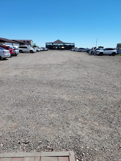 The image shows a wide-angle view of a gravel parking lot on a clear, sunny day. Several vehicles are parked in rows, and a large, open-air building with a dark roof and glass walls is visible in the background. The building appears to be a restaurant or event venue, with people seated at tables inside. A few people are standing near a white SUV in the mid-ground. The foreground consists of the textured gravel surface of the parking lot, with a concrete curb and brick paving at the very bottom edge of the image. The sky is a solid, bright blue. The location is identified as Toranghylyk, Kazakhstan.