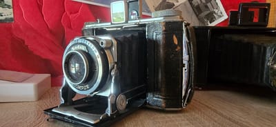The image shows a vintage Kodak camera with a bellows, placed on a wooden surface. The camera is black with silver accents and has the brand name "Kodak" and "COMPUR" visible on the lens assembly. A red, textured background is behind the camera, with some black and white photographs or prints visible. A white object, possibly a camera accessory or case, is to the left of the camera. The scene is indoors, likely on a table or shelf. There are no people or animals present. The lighting suggests a casual indoor setting. There is no visible indication of the location being in Gostagayevskaya, Russia.