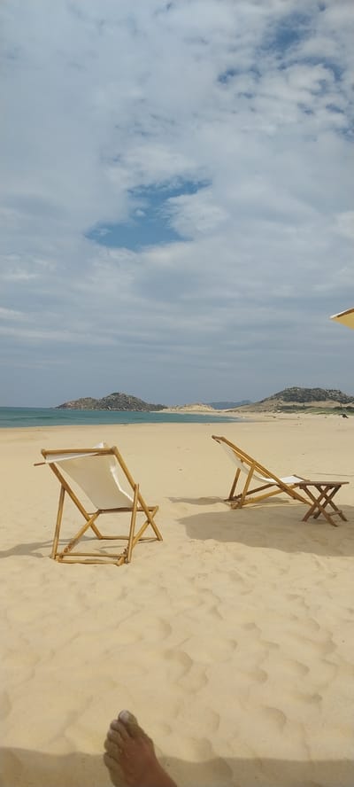 In Cát Tiến, Vietnam, an image captures a tranquil beach scene under a cloudy sky. Two empty, light-colored canvas deck chairs with bamboo frames are positioned on the golden sand. The chair on the left is closer to the foreground and angled slightly away from the viewer, while the chair on the right is further back and positioned to the right of the frame. A small, wooden X-shaped table sits next to the right chair. In the distance, the turquoise ocean meets the horizon, with two rocky, tree-covered islands visible on the left side of the frame. The beach itself shows subtle ripples in the sand. The bottom left corner of the image shows a person's foot, covered in sand, suggesting they are relaxing on the beach. A small portion of a yellow umbrella is visible at the top right edge of the image.